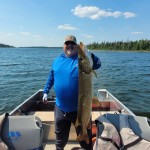 Person on a boat holding a large fish with one hand, standing under a clear blue sky on a lake with trees in the background.