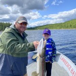 Two people standing on a boat by a lake. The adult is holding a large fish, and both are smiling. Trees line the shore.