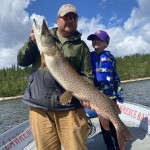A person holds a large fish on a boat, with a child beside them, against a backdrop of water and trees under a partly cloudy sky.