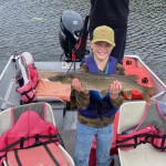 Child smiling while holding a fish on a small boat with life jackets and motor, near another person facing away.