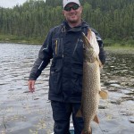 Man in a blue fishing outfit holding a large fish on a boat in a lake, with a forested shoreline in the background.