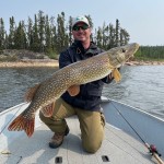 Person on a boat, holding a large fish with a smile. Forested shoreline and calm water visible in the background.