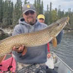 Man in a boat holding a large fish, with another person partially visible in the background; trees and lake in the background.