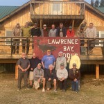 Group of people posing in front of a wooden building with a "Lawrence Bay Lodge" sign.