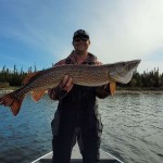 Person on a boat holding a large fish horizontally with both hands. Water and trees are in the background.
