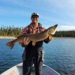 Person stands on a boat holding a large fish, with trees and water in the background under a clear sky.