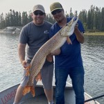 Two men on a boat hold a large fish, with trees and water in the background.