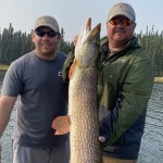 Two men standing on a boat hold a large fish between them, with a lake and forest in the background.