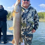 Person proudly holding a large fish on a boat, wearing camo clothing and sunglasses, with a forested shoreline in the background.
