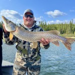 Person in a camo jacket and cap holding a large fish on a boat, with a lake and forested area in the background.