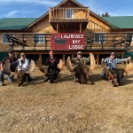 Four people kneel in front of a lodge with moose antlers and hides on the ground in front of them.
