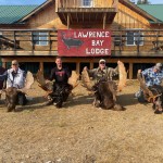 Five people kneel beside moose antlers in front of Lawrence Bay Lodge, with a log building in the background.