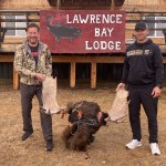 Two men holding large antlers stand in front of a sign reading "Lawrence Bay Lodge" with a moose carcass between them.