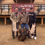 Three men stand outside holding moose antlers, with a moose head on the ground. Wooden building in the background with a sign.