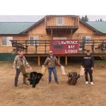 Three people stand in front of Lawrence Bay Lodge holding moose antlers and a rack on the ground.