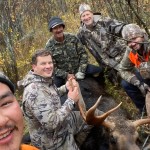 Four hunters in camouflage pose with a moose in a forested area. Text reads, "Moose number 7!.