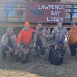 Five people kneel next to two moose at Lawrence Bay Lodge, with a wooden sign above them.
