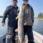 Two people on a boat holding a large fish, one wearing sunglasses and a cap, on a sunny day with water and trees in the background.