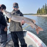 Person on a boat holding a large fish they caught, wearing a cap and sunglasses, with another person standing nearby.