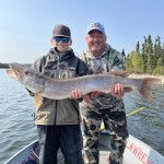 Two men on a boat holding a large fish they caught, with trees and a lake in the background.