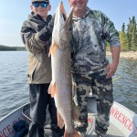 Two people on a boat hold a large fish, smiling. Trees and water are visible in the background.