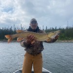 Person standing on a boat holding a large fish with both hands. Trees and cloudy sky in the background.