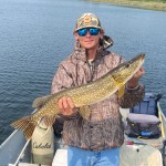 Person holding a large fish on a boat, wearing camouflage clothing, sunglasses, and a cap, with a lake in the background.