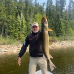 Man on a boat holding a large fish vertically with a forest backdrop. He is wearing a cap, a jacket, and khaki pants.