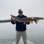 Person standing on a boat holding a large fish with both hands, lake and trees in the background.