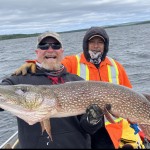 Two men on a boat hold a large fish. One wears camouflage, gloves, and sunglasses. The other wears an orange jacket.