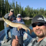 Three people on a boat, one holding a large fish, with a forested shoreline in the background, all giving thumbs-up.