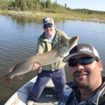 Two men on a boat in a lake. One holds a large fish, while the other takes a selfie. Trees line the distant shore.