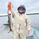 Person in rain gear holding a large fish on a boat in a lake under an overcast sky.
