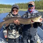 Two people on a boat hold a large northern pike fish in front of a forested shoreline.