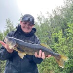 Person in a black jacket and cap holding a large fish outdoors with trees in the background.
