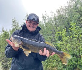 Person in a black jacket and cap holding a large fish outdoors with trees in the background.