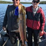 Two people on a boat hold up a large fish they caught, with a lake and forested shoreline in the background.