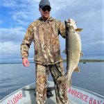 Person in camouflage clothing stands on a boat holding a large fish, with water and sky in the background.