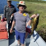 Two men in a boat by a river; one man holds a large fish while the other stands nearby.