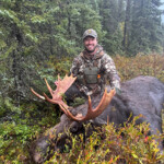 A man in camouflage kneels beside a large moose lying on the ground in a forested area.