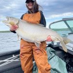 Person wearing orange overalls holds a large fish on a boat with water and a cloudy sky in the background.