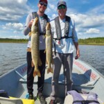 Two men standing on a boat each holding a large fish, with a lake and trees in the background.