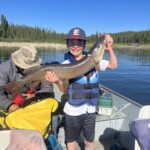 Smiling boy in life jacket holds up a large fish on a boat, with another person seated beside him and a lake in the background.