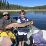 A boy in a life jacket holds a large fish on a boat, with an adult seated beside him on a lake with forested shoreline.