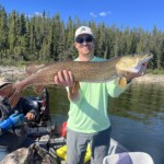 Man standing on a boat holding a large northern pike fish with a forested shoreline in the background.