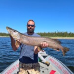 Man standing on a boat holding a large fish with both hands, with water and trees visible in the background.