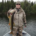 Man in camouflage clothing stands on a boat holding a large fish, with trees and water in the background.