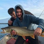 Two men on a boat; one holds a large fish while the other gestures with a knife near his face.