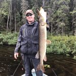 Man in dark outdoor clothing standing on a boat holding a large fish upright with trees and greenery in the background.