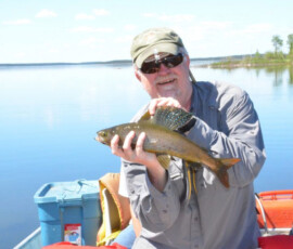 Man wearing sunglasses and a cap holding a fish on a boat with a lake and trees in the background.